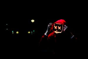 Eerie digital face with X's for eyes beneath a red hood, with hands about to draw the hood away, all against a dark nightime background