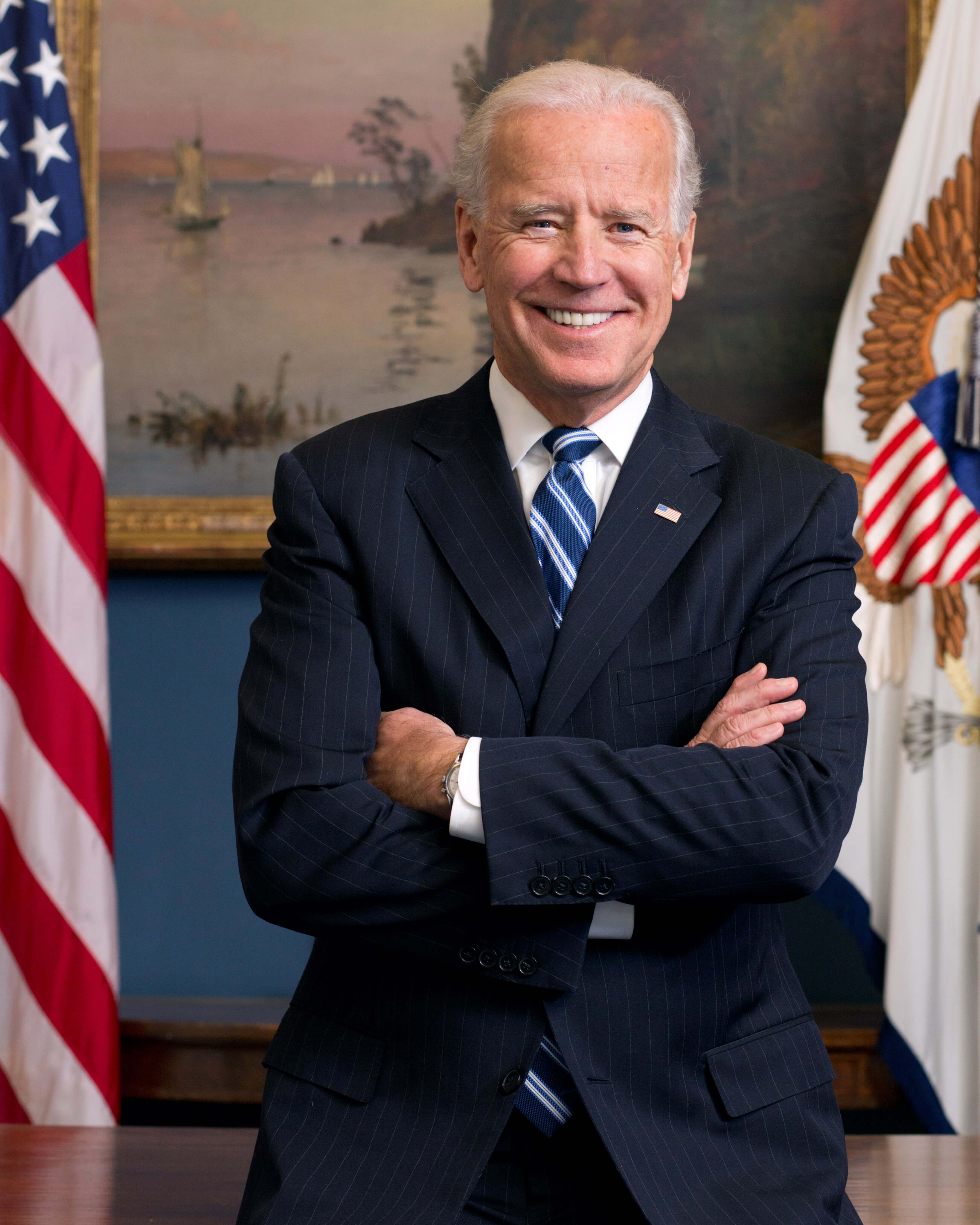 Official portrait photo of Joe Biden, arms crossed in a dark blue suit as he leans against a desk with a US flag to his right