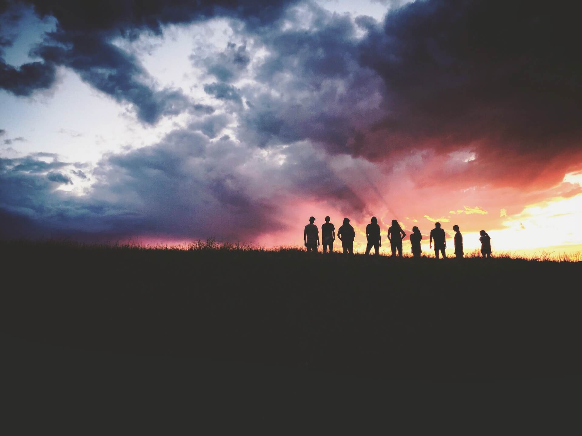 Image of nine distant people in silhouette with the ground in front of them long and dark and the horizon behind them half stormy and half sunset