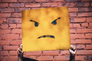 A square, yellow metal sign with signs of rust and a frowny face emoji on it, held aloft by two hands in front of a brick wall