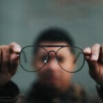 Blurred image of man's face behind clear foreground image of his glasses held in his hands far out from his face