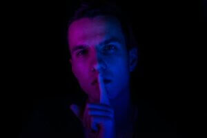 White man sushing the viewer with finger to lips, illuminated in purple and blue against a dark background