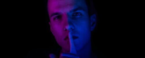 White man sushing the viewer with finger to lips, illuminated in purple and blue against a dark background