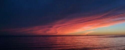 The sea at sunset, with the upper half of the image being dark clouds illuminated in reds and oranges only at the bottom with only a hint of clear sky at the horizon and the lower half calm water, barely illuminated