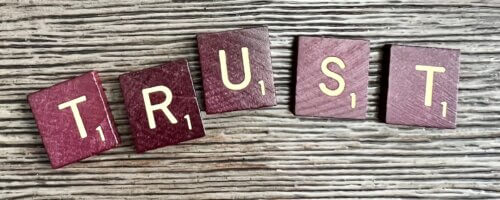 Five reddish-brown Scrabble tiles spelling out "trust" atop an ash-brown woodgrain surface