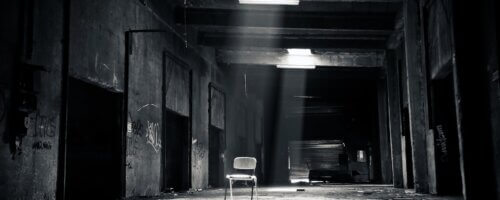 Black and white image of a spooky, mostly empty warehouse-like room with an empty chair just off-center and two openings in the roof letting in weak shafts of light