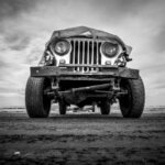 black-and-white image of the front of a slightly busted-up old jeep, taken from ground level looking up, with a cloudy sky in the background