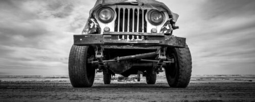 black-and-white image of the front of a slightly busted-up old jeep, taken from ground level looking up, with a cloudy sky in the background