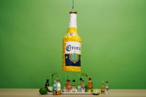 A big beer-bottle shaped pinata hanging over a bunch of small bottles of liquor in front of a green backgroun