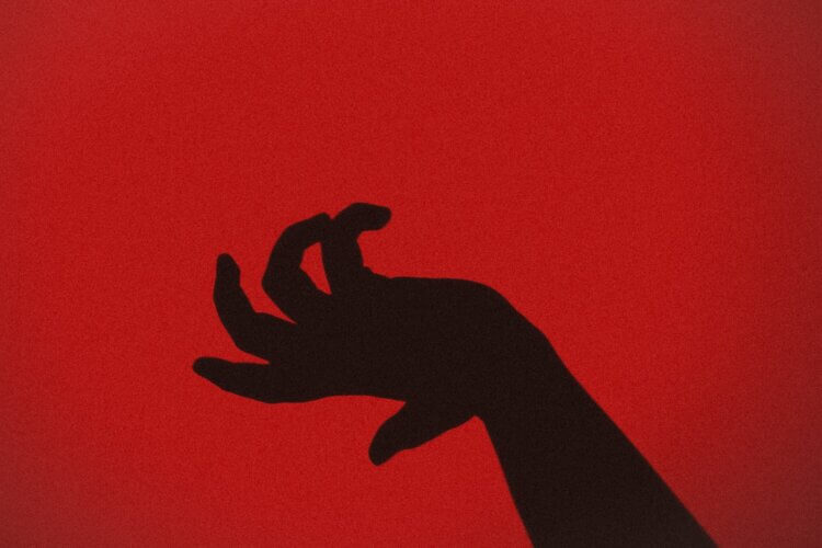 A silhouetted black hand in front of a dark red background, open and fingers curled as if to slap or grab something