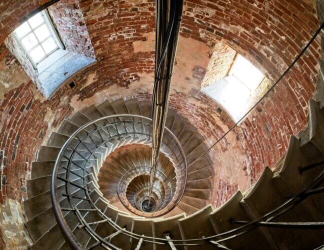overhead view of a spiral staircase in a round brick tower descending downward, with light coming in from small windows on either side