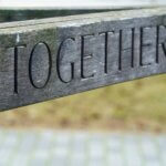A grayish piece of wood, either a narrow sign or part of a fence, with the word "TOGETHER" carved in black into it