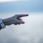 A blurry white and blue background, possibly the sky and water, with a white person's arm wearing a white watch extending from the left-hand side, angled upward and the index finger pointing aggressively toward the right of the image
