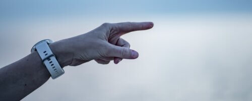 A blurry white and blue background, possibly the sky and water, with a white person's arm wearing a white watch extending from the left-hand side, angled upward and the index finger pointing aggressively toward the right of the image