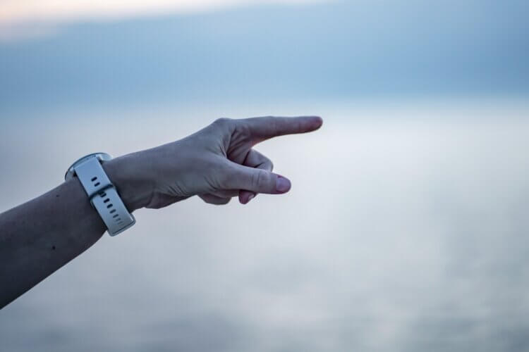 A blurry white and blue background, possibly the sky and water, with a white person's arm wearing a white watch extending from the left-hand side, angled upward and the index finger pointing aggressively toward the right of the image