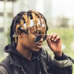 Black man with brown and blond locs lowering his glasses down his nose and looking out over the top of them