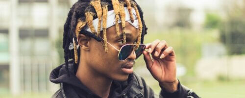 Black man with brown and blond locs lowering his glasses down his nose and looking out over the top of them