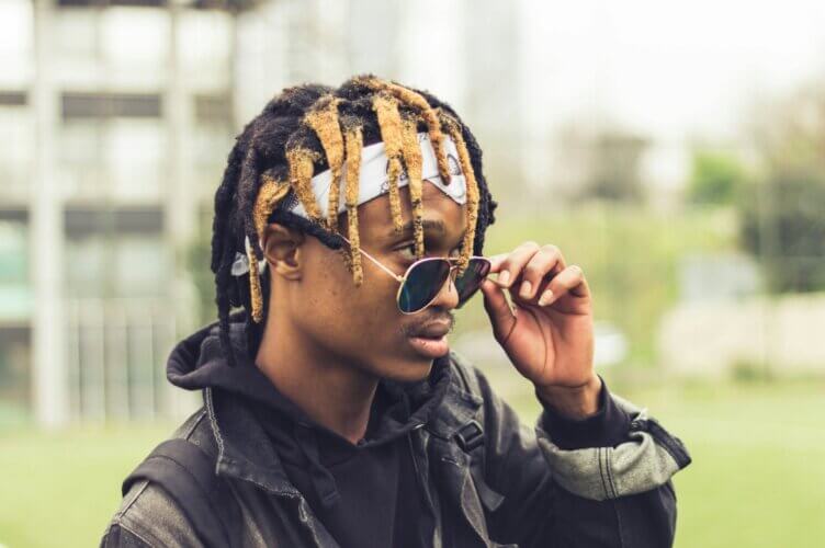Black man with brown and blond locs lowering his glasses down his nose and looking out over the top of them