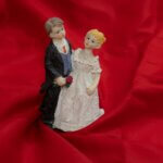 A "cutesy" figurine set of a white bride and groom (each looking almost childlike or cherubish), possibly a cake-topper, against a background of red silk
