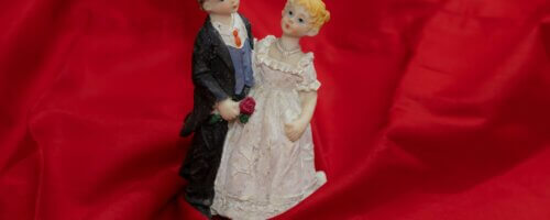 A "cutesy" figurine set of a white bride and groom (each looking almost childlike or cherubish), possibly a cake-topper, against a background of red silk