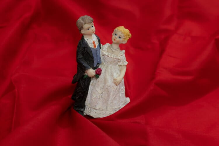 A "cutesy" figurine set of a white bride and groom (each looking almost childlike or cherubish), possibly a cake-topper, against a background of red silk