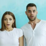 A female-and-male couple, both in white t-shirts, standing in front of a light blue background with shocked/surprised expressions on their faces