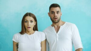 A female-and-male couple, both in white t-shirts, standing in front of a light blue background with shocked/surprised expressions on their faces