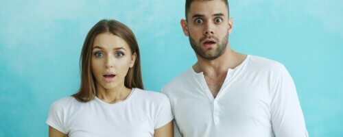 A female-and-male couple, both in white t-shirts, standing in front of a light blue background with shocked/surprised expressions on their faces