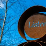 Image of a weathers and oxidized artistic display with the word "listen" craved through the metal disc at the center of the main disk of the display, the blue sky behind showing through that carved out word