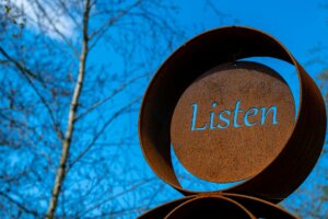 Image of a weathers and oxidized artistic display with the word "listen" craved through the metal disc at the center of the main disk of the display, the blue sky behind showing through that carved out word