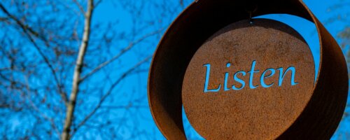 Image of a weathers and oxidized artistic display with the word "listen" craved through the metal disc at the center of the main disk of the display, the blue sky behind showing through that carved out word