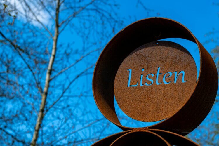 Image of a weathers and oxidized artistic display with the word "listen" craved through the metal disc at the center of the main disk of the display, the blue sky behind showing through that carved out word