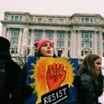 Woman in a pink knit cap holding a colorful sign that pictures a raised fist and the word "RESIST"