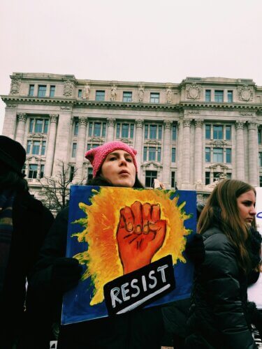 Woman in a pink knit cap holding a colorful sign that pictures a raised fist and the word "RESIST"