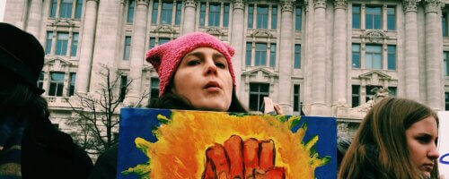 Woman in a pink knit cap holding a colorful sign that pictures a raised fist and the word "RESIST"