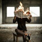 Man sitting cross-legged on an old wooden table holding a burning newspaper in front of his face in an abandoned concrete room