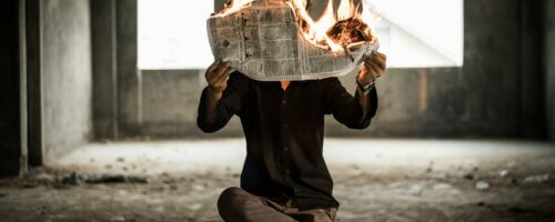 Man sitting cross-legged on an old wooden table holding a burning newspaper in front of his face in an abandoned concrete room