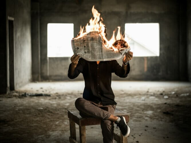 Man sitting cross-legged on an old wooden table holding a burning newspaper in front of his face in an abandoned concrete room