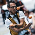Man with glasses in white shirt, black-and-gold vest and juggling three orange balls