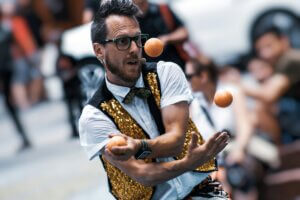 Man with glasses in white shirt, black-and-gold vest and juggling three orange balls
