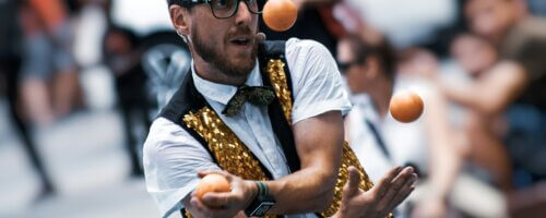 Man with glasses in white shirt, black-and-gold vest and juggling three orange balls