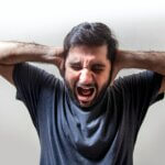 A man with black hair and close-cropped beards wearing a blue T-shit, holding his hands to the side of his head and crying out with a grimaced expression