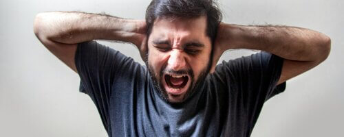 A man with black hair and close-cropped beards wearing a blue T-shit, holding his hands to the side of his head and crying out with a grimaced expression