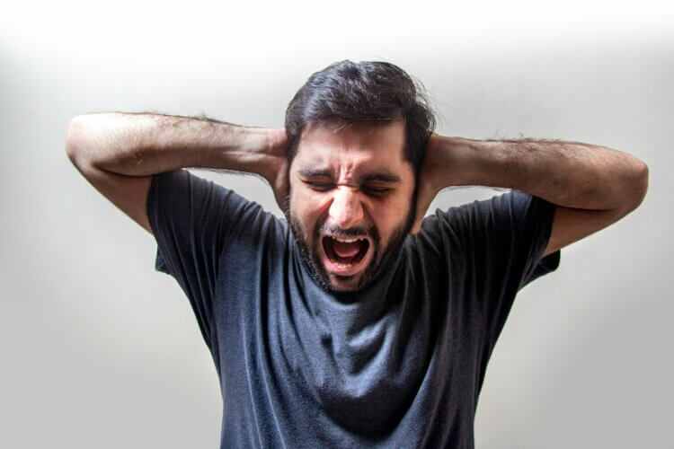 A man with black hair and close-cropped beards wearing a blue T-shit, holding his hands to the side of his head and crying out with a grimaced expression