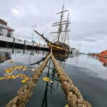image of an old-style sailing vessel from a distance at pier, with two mooring ropes extending from near view to the ship in the distance
