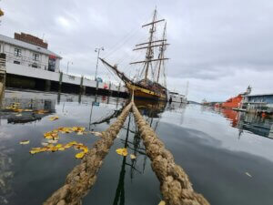 image of an old-style sailing vessel from a distance at pier, with two mooring ropes extending from near view to the ship in the distance