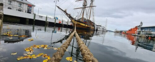image of an old-style sailing vessel from a distance at pier, with two mooring ropes extending from near view to the ship in the distance