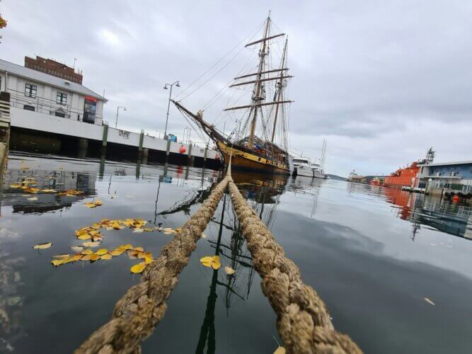 image of an old-style sailing vessel from a distance at pier, with two mooring ropes extending from near view to the ship in the distance