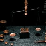 A set of copper or bronze balance-style scales with one end higher than the other, on a dark tabletop with small cups and other paraphernalia scattered about