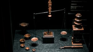 A set of copper or bronze balance-style scales with one end higher than the other, on a dark tabletop with small cups and other paraphernalia scattered about